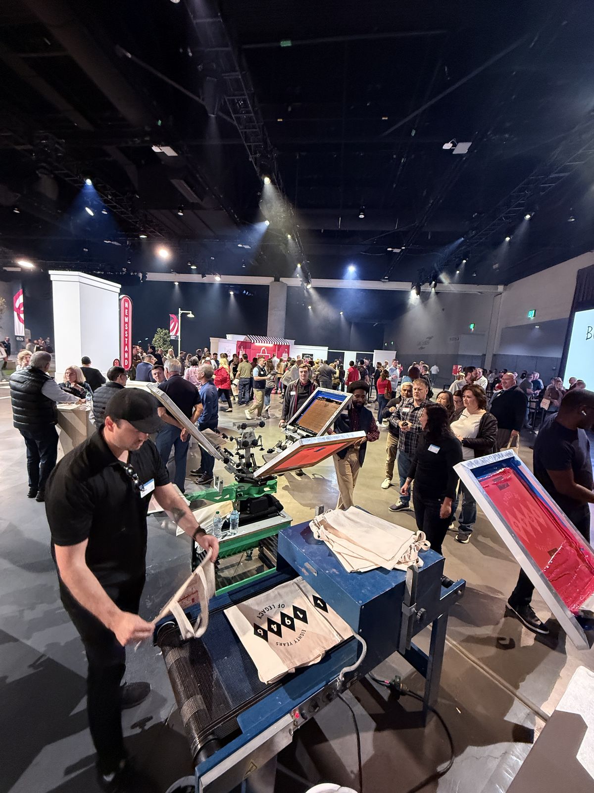 Convention hall crowd around a live Merch Troop station with anniversary totes moving through a dryer